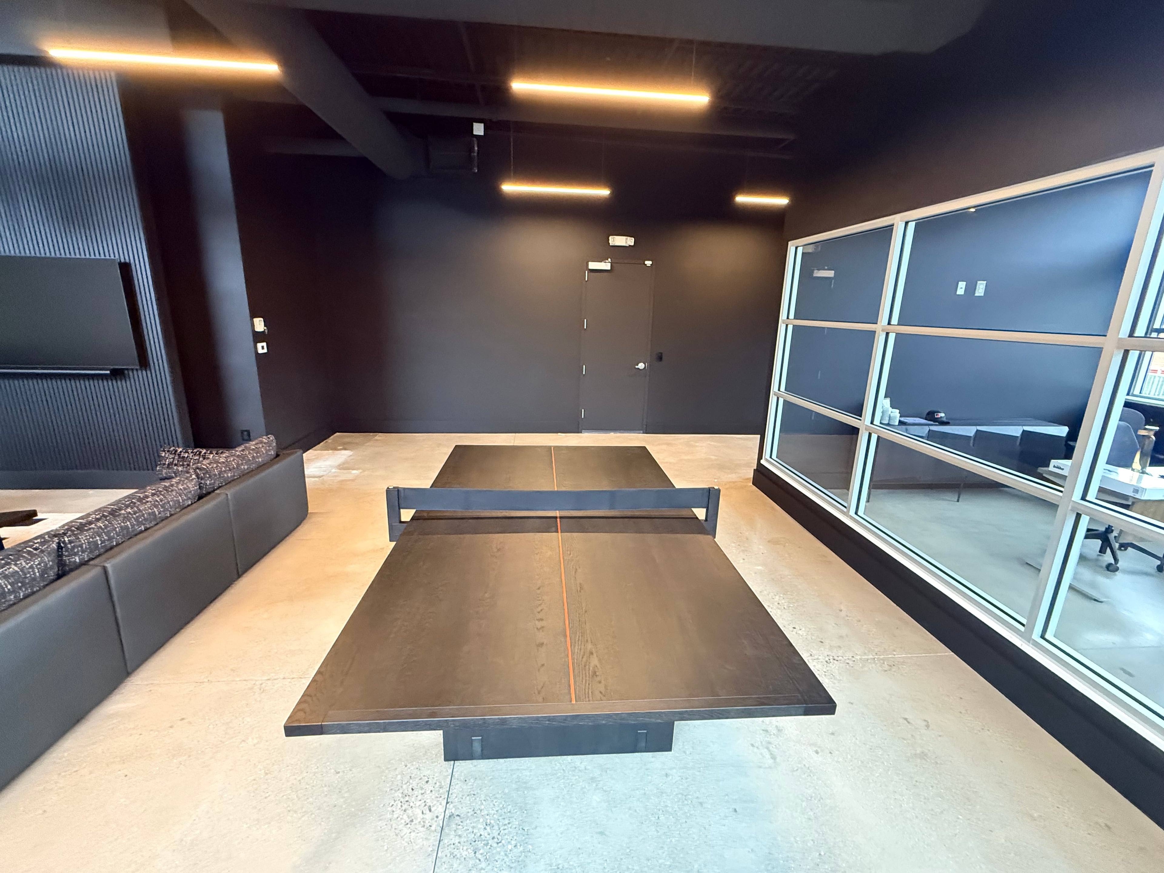 Completed corporate entertainment lounge with matte black walls, polished concrete floor, ping-pong table, gray sectional sofa, wall-mounted TV, floor-to-ceiling glass office partition wall, and linear LED ceiling lighting: commercial build-out by JD Contracting LLC
