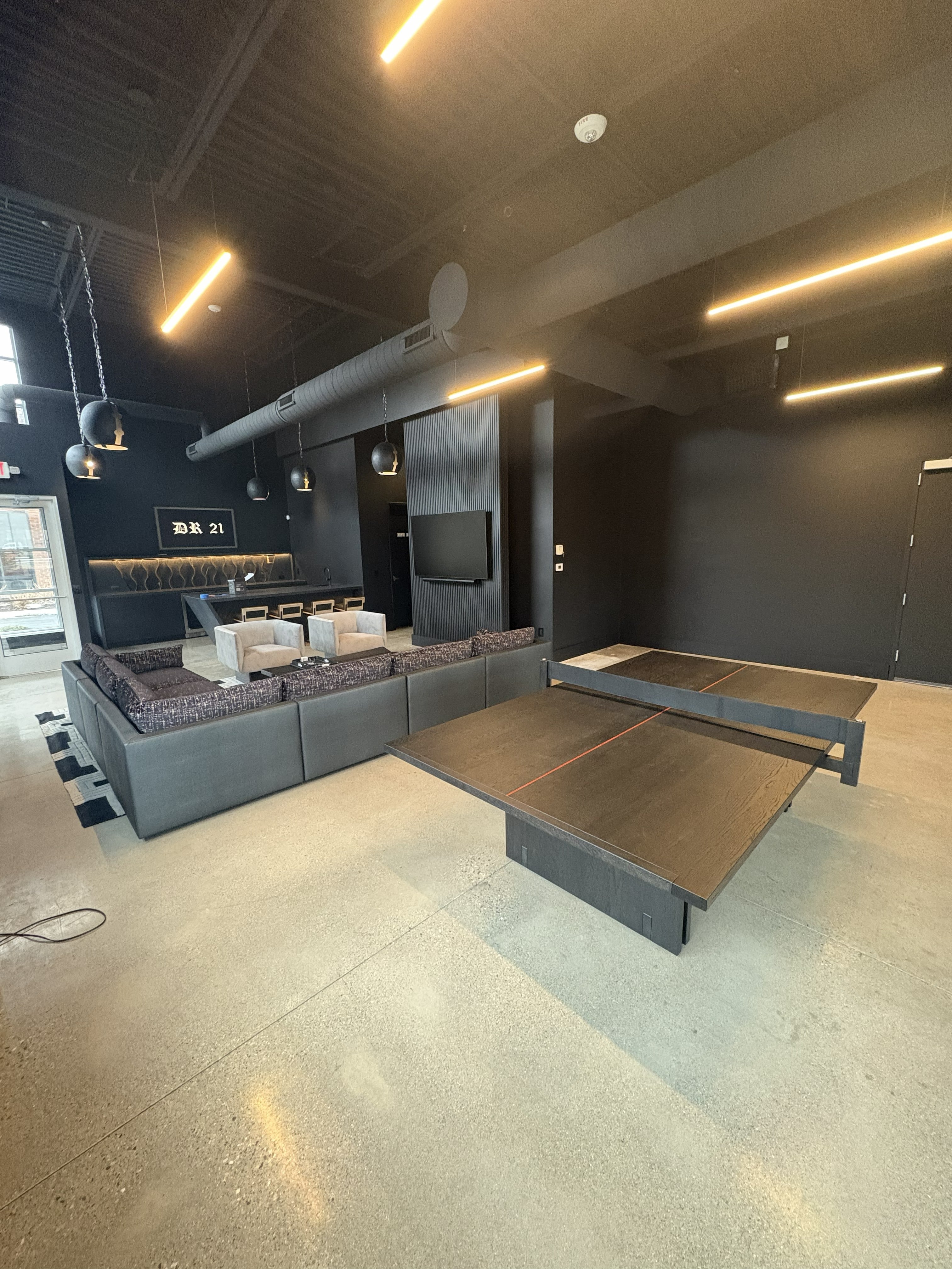 Corporate entertainment lounge build-out with all-black walls and exposed industrial ceiling, large gray sectional sofa, ping-pong table, polished concrete floor, bar area with high-top seating and DR 21 signage, globe pendant lights, and linear LED strip lighting: built by JD Contracting LLC