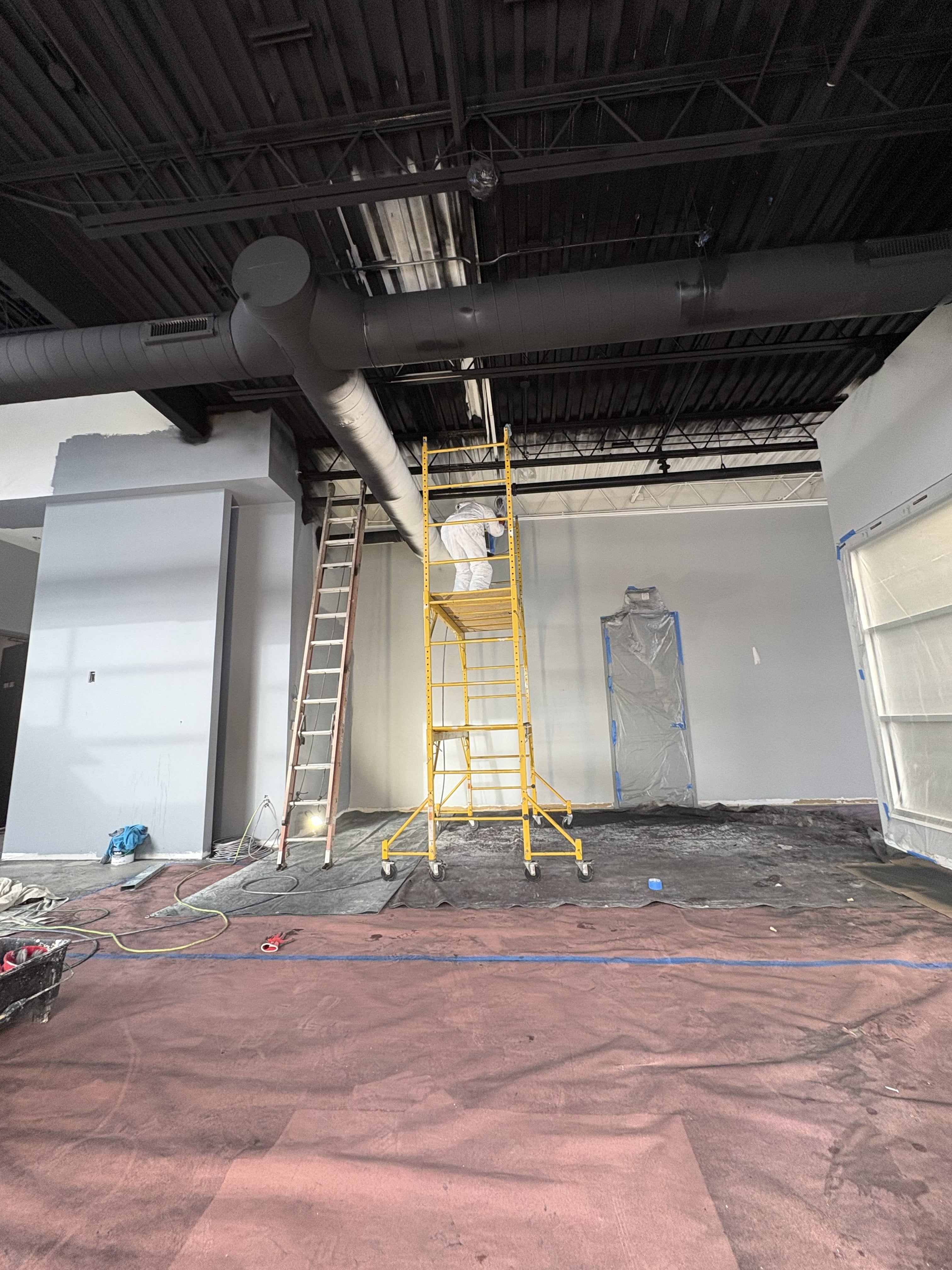 Commercial tenant build-out in progress with painter on scaffold tower finishing gray drywall partitions, exposed black steel bar joist ceiling, large-diameter HVAC ductwork, floor protection, and active construction equipment: by JD Contracting LLC