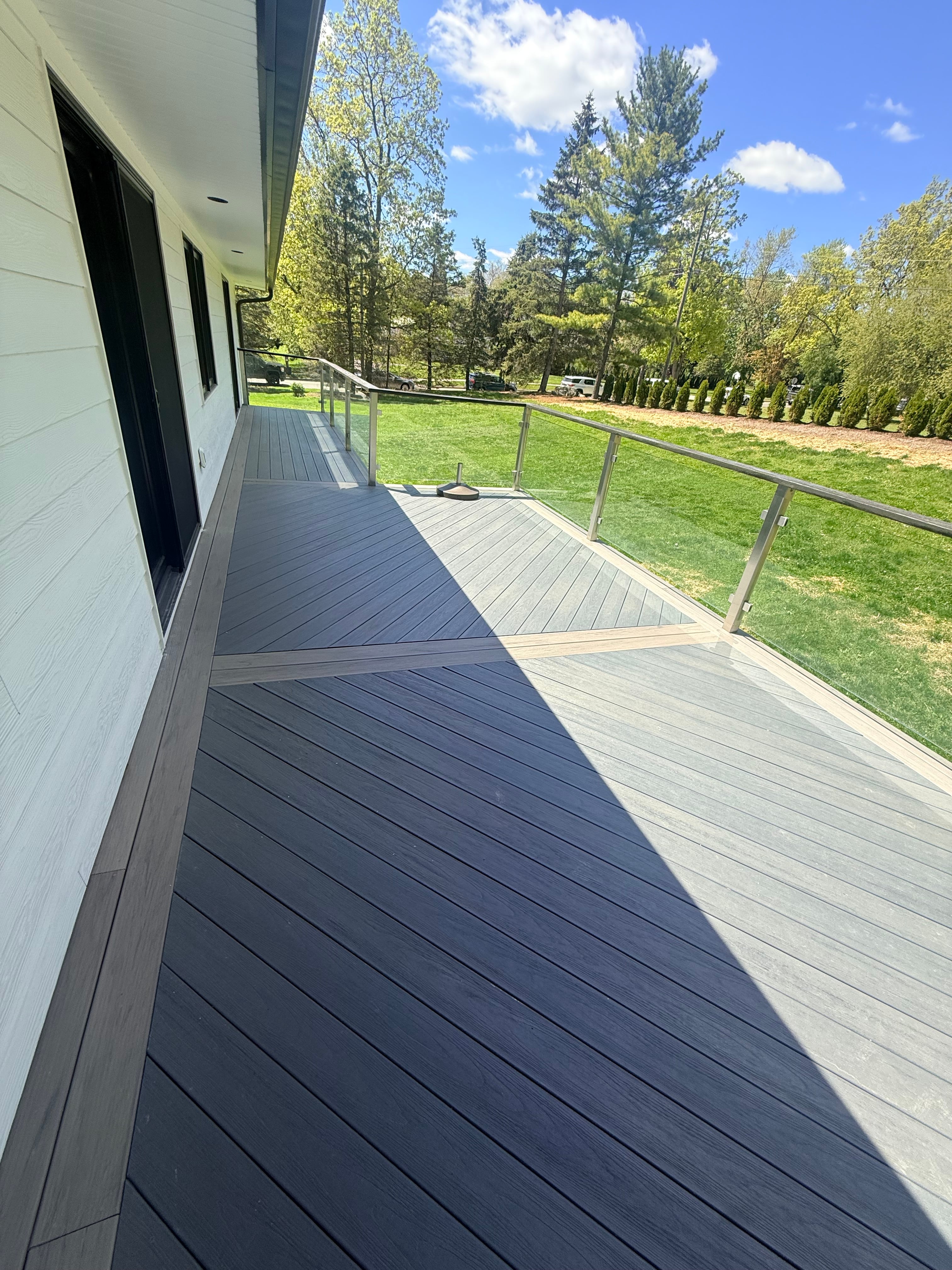 Dark charcoal composite deck with diagonal board pattern, light gray picture-frame border, and cable wire railing: multi-level installation with steps leading to lower tier, lush green backyard in background, built by JD Contracting LLC