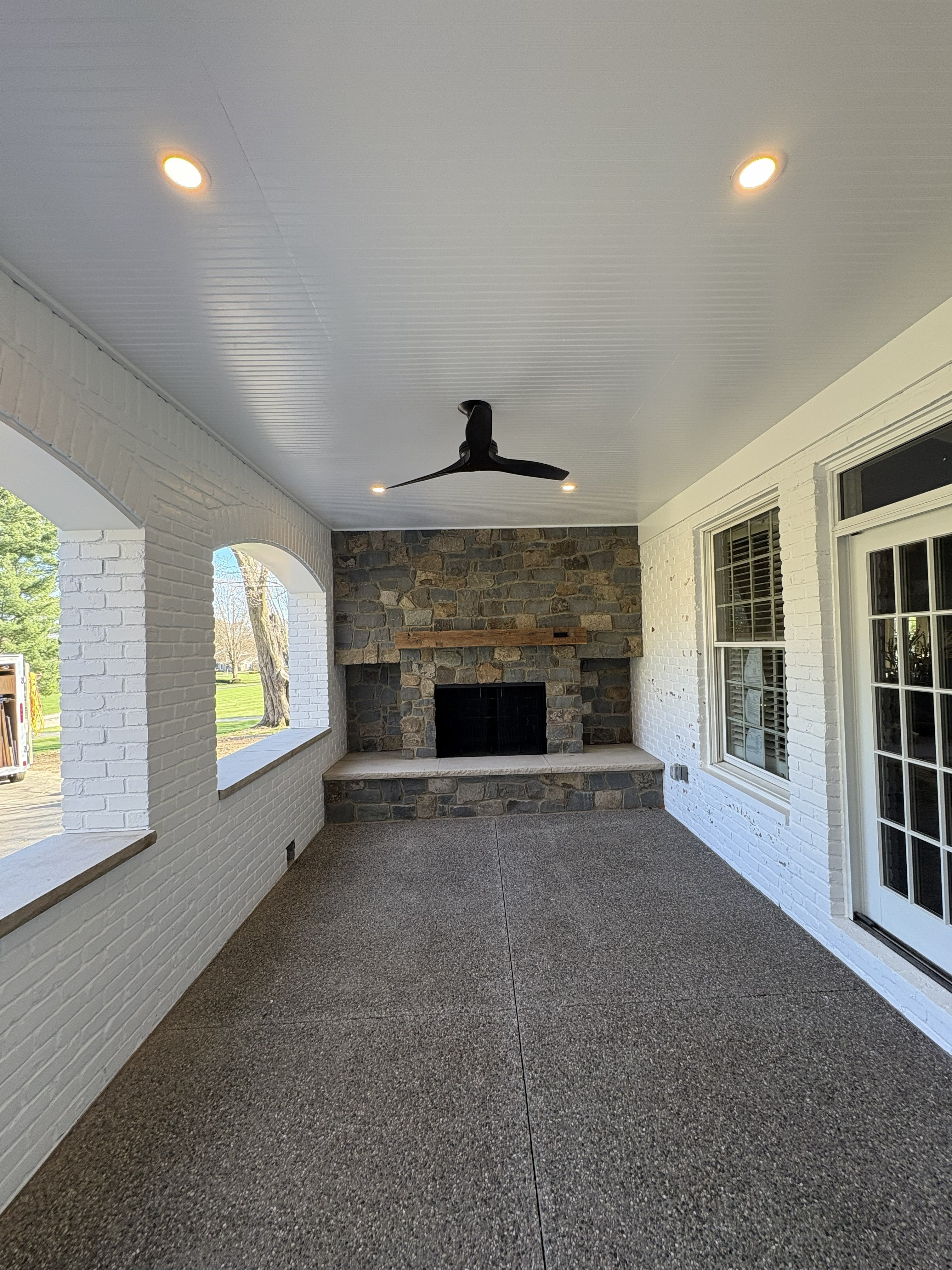 Covered outdoor living space with floor-to-ceiling stacked stone fireplace, white beadboard ceiling with recessed lighting and ceiling fan, and painted brick walls: built by JD Contracting LLC