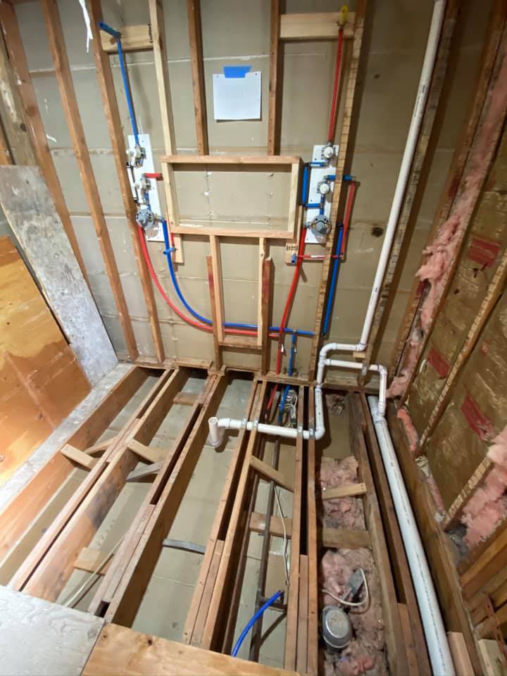 Rough plumbing installation showing blue PEX cold and red PEX hot supply lines, shower valve rough-ins, and PVC drain pipes through wood stud wall framing with fiberglass insulation: residential plumbing by JD Contracting LLC