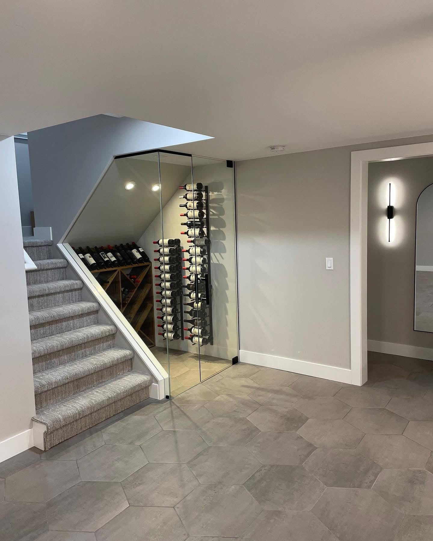 Finished basement with glass-enclosed wine cellar built under carpeted staircase displaying floor-to-ceiling wine bottle racks, large hexagonal gray tile floor, smooth gray painted walls, arched doorway with round mirror and modern wall sconce, and recessed lighting throughout: by JD Contracting LLC