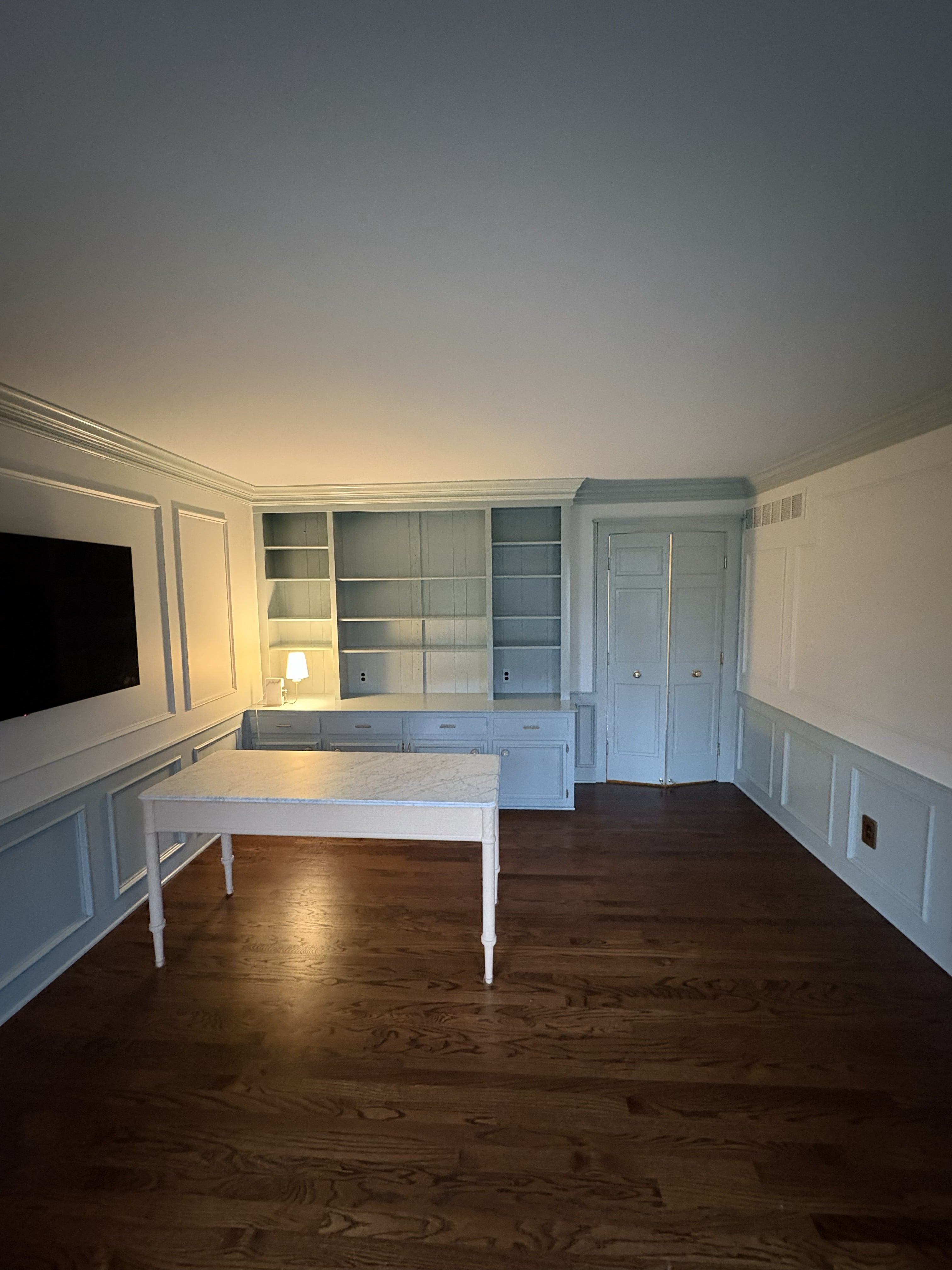 Custom built-in home office with pale blue-gray floor-to-ceiling shelving unit, lower cabinet drawers with silver bar pulls, integrated door, TV inset in picture-frame panel wall, matching wainscoting throughout, white marble-top desk, and dark red oak hardwood floors: by JD Contracting LLC