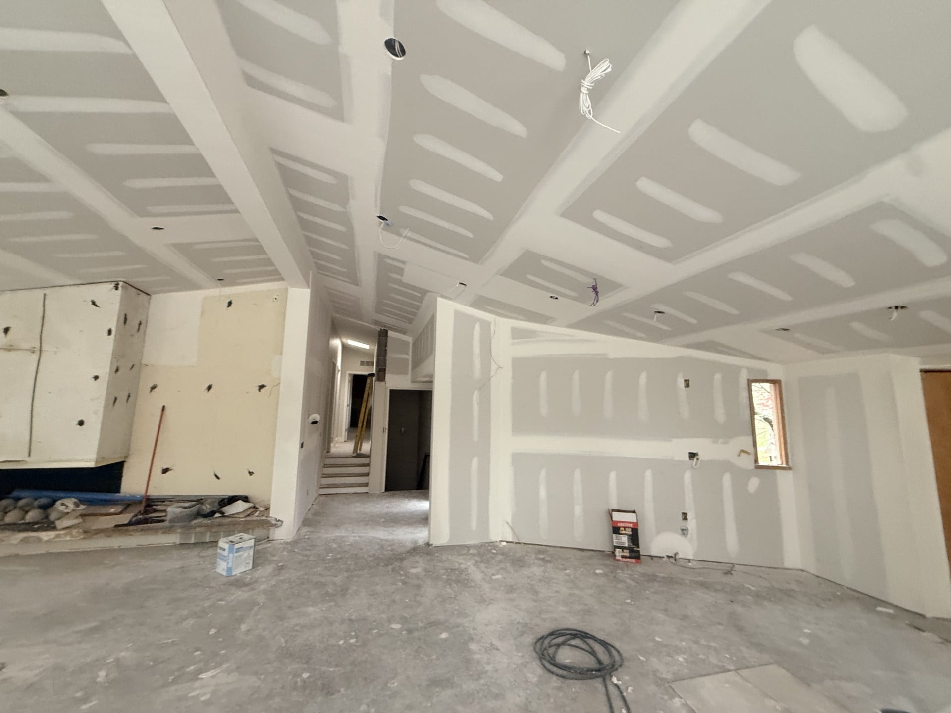 Whole-home drywall installation in progress showing freshly hung drywall with taped seams and joint compound on walls and a coffered ceiling, recessed light cutouts, and active construction site: by JD Contracting LLC