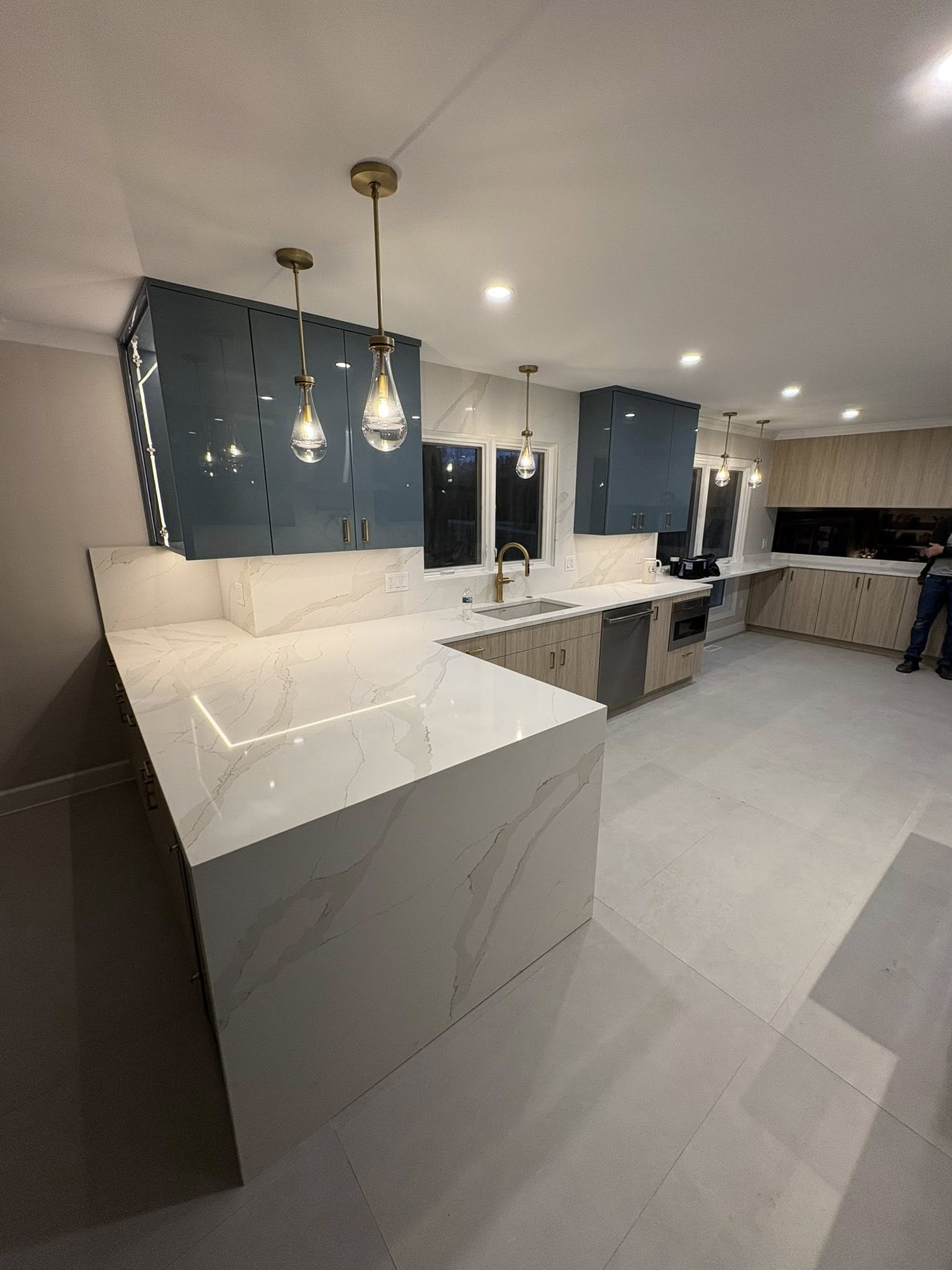 Large kitchen with steel blue high-gloss upper cabinet doors with brass hardware, light natural wood lower cabinets, waterfall Calacatta quartz peninsula, teardrop glass pendant lights, and large-format tile floor: custom cabinetry by JD Contracting LLC