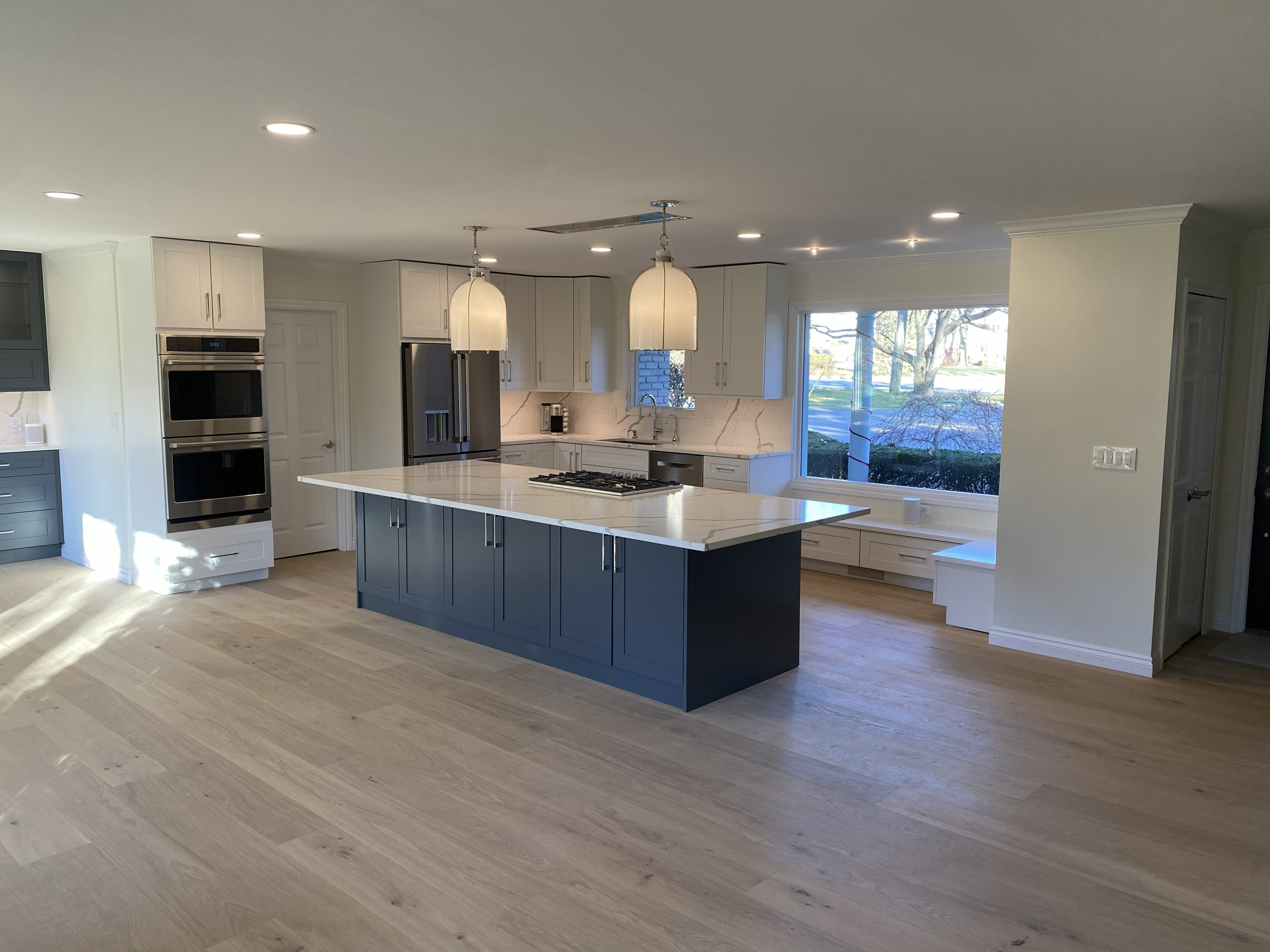Open-concept kitchen remodel with navy blue shaker center island, white shaker upper cabinets, waterfall quartz countertops, double stainless wall ovens, french door refrigerator, dome pendant lights, light oak hardwood floors, and built-in garden window seat: by JD Contracting LLC